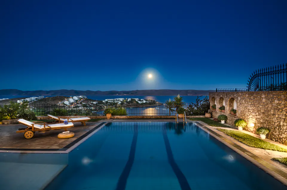 Swimming pool at night with sun loungers, and the moon over the bay
