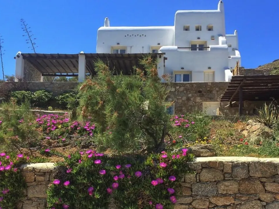 Λευκή βίλα πίσω από πέτρινο τοιχίο και ροζ λουλούδια