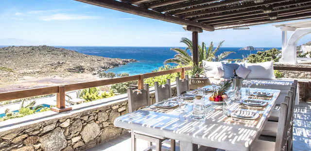 A table laid on a terrace with a wooden pergola, looking over a bay and the sea